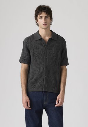 Mand iført mørkegrå kortærmet strikket skjorte med knapper og teksturerede geometriske mønstre samt mørkeblå jeans, set forfra mod en ensfarvet baggrund.