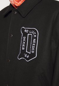 Black wool blend jacket featuring a prominent embroidered patch with “NO LUCK NEEDED” and "DICKIES '22" in white text. Fastened with black buttons.