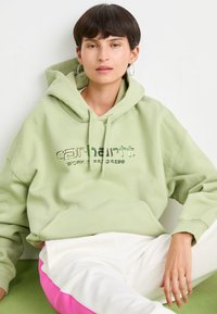 Ungdom med kort, mørkt hår som har på seg hoop-øredobber, en lys grønn Carhartt hettegenser og hvite bukser med rosa striper, som sitter mot en hvit vegg.