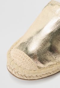 Metallista kultahappo yläosa, jossa on kudottu beige kangaspistokas. Punottu jutepinta pohjan ympärillä tarjoaa tekstuurista jatkuvuutta materiaalien välillä.