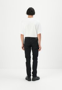 Black jeans with a straight fit and back pockets, paired with a loose-fitting white t-shirt, viewed from the back. Black shoes complete the look.