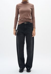 Femme portant un pull roulé marron, un jean noir ample avec une ceinture, des mocassins noirs, et tenant une pochette noire dans sa main gauche.