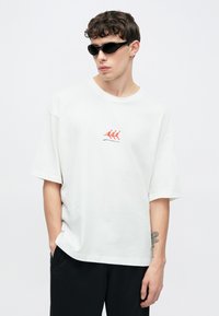 T-shirt blanc oversize à manches courtes, avec un motif brodé rouge représentant trois figures. Accessoirisé avec des lunettes de soleil noires. Pantalon noir.