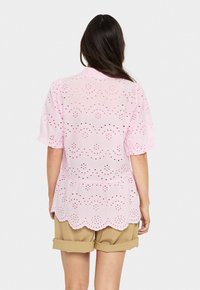 Rosa Augenbluse mit gewellten Saum, kurzen Puffärmeln und durchbrochenem Blumenmuster, getragen über beigefarbenen, gerollten Shorts.
