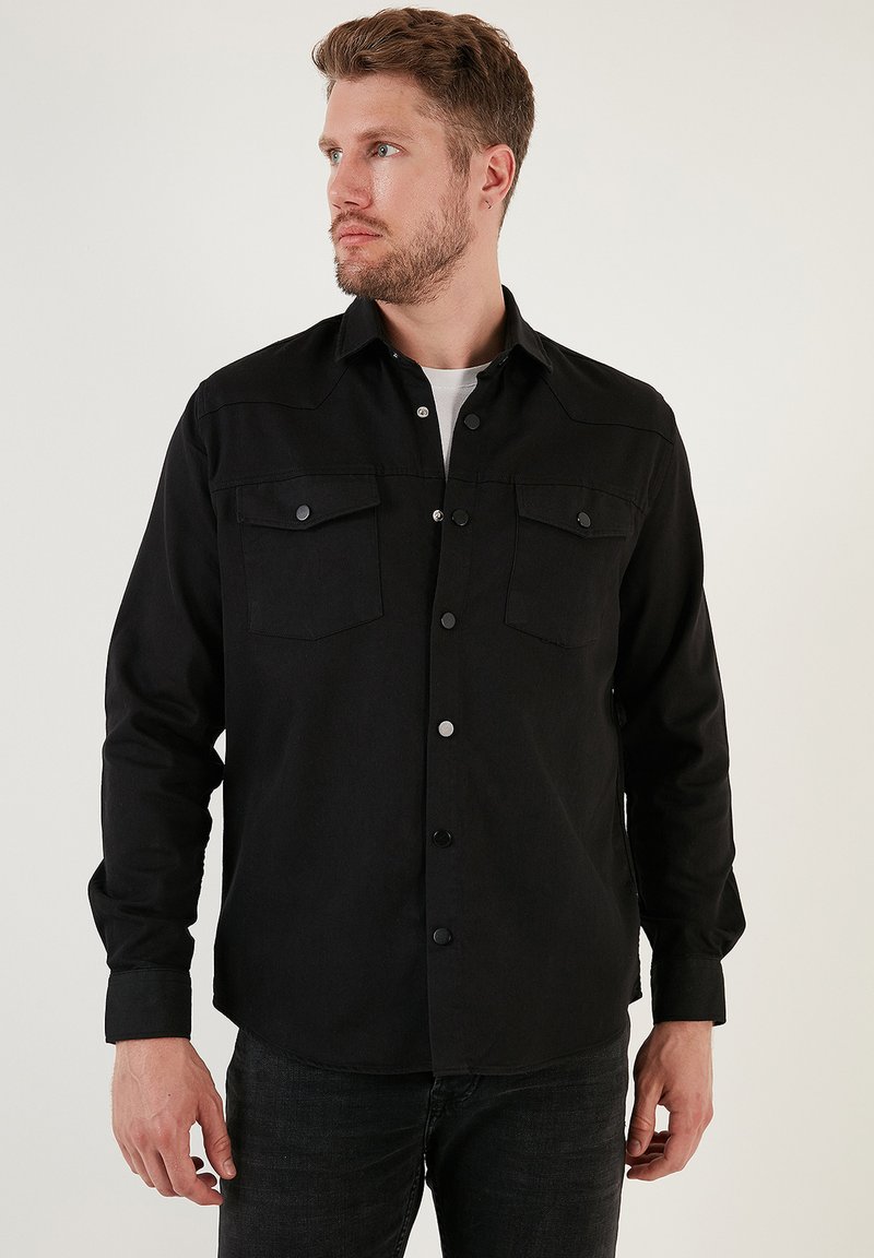 Camicia nera con bottoni in cotone misto, dotata di due tasche sul petto, bottoni a scatto e colletto classico. Texture liscia e design aderente.