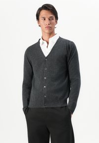 Uomo che indossa un cardigan grigio scuro con bottoni sopra una camicia bianca con colletto e pantaloni neri, in piedi con le mani in tasca contro uno sfondo bianco.