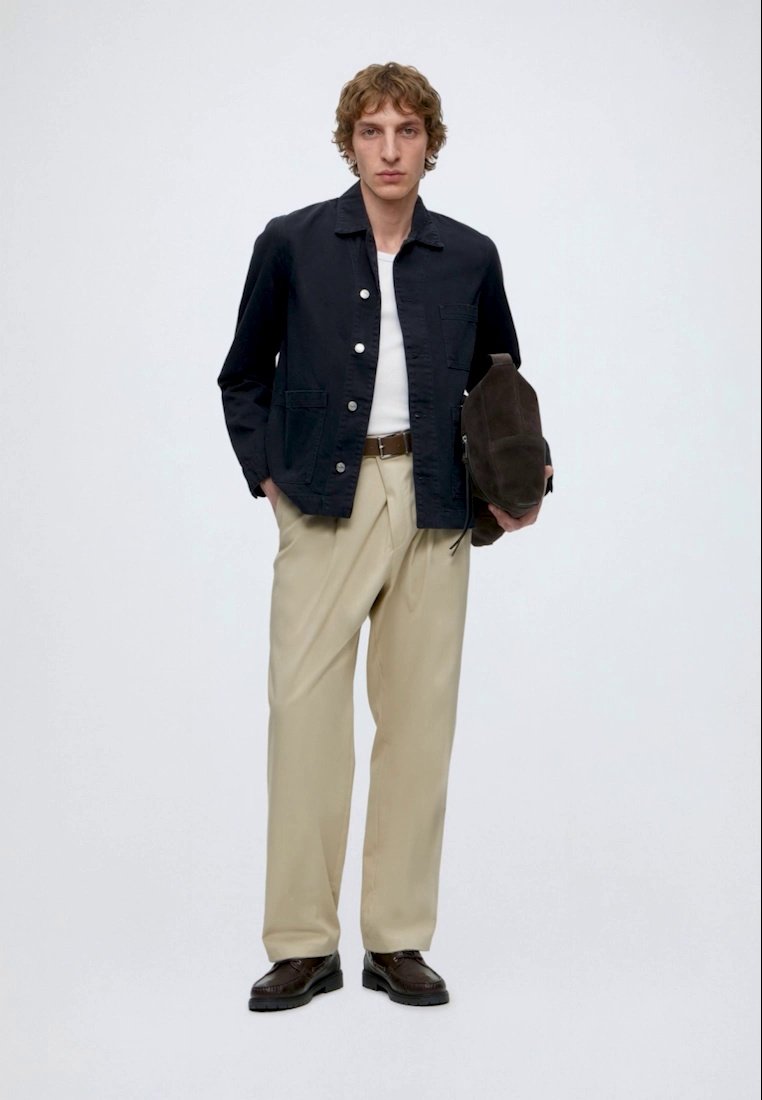 Homme debout de face, portant une veste bleu marine, une chemise blanche, un pantalon beige, des chaussures foncées et tenant un sac marron sur un fond uni.