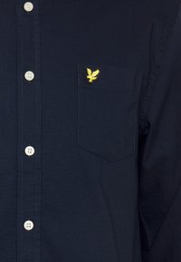 Camisa azul-marinho de manga longa, feita de tecido suave, com um bolso no peito e um logótipo de águia bordado em amarelo no bolso.