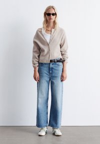 Beige Strickjacke mit Knöpfen, weißes Tanktop, hellblaue weite Jeans und weiße Sneakers mit grünen Akzenten, getragen mit schwarzen Sonnenbrillen.
