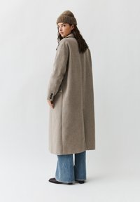 Vrouw die een beige lange jas draagt, een bruin gebreide muts, blauwe wijde broekspijpen en zwarte schoenen, staand en over haar schouder kijkend.