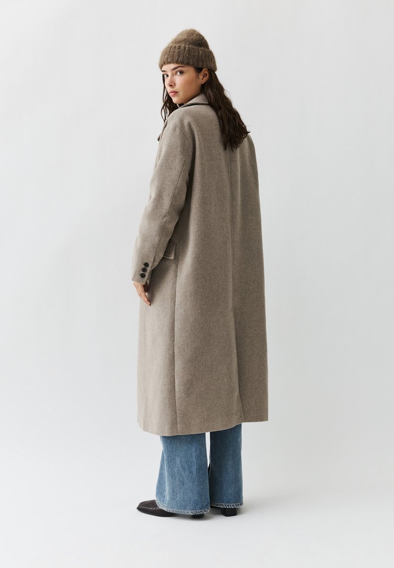 Vrouw die een beige lange jas draagt, een bruin gebreide muts, blauwe wijde broekspijpen en zwarte schoenen, staand en over haar schouder kijkend.