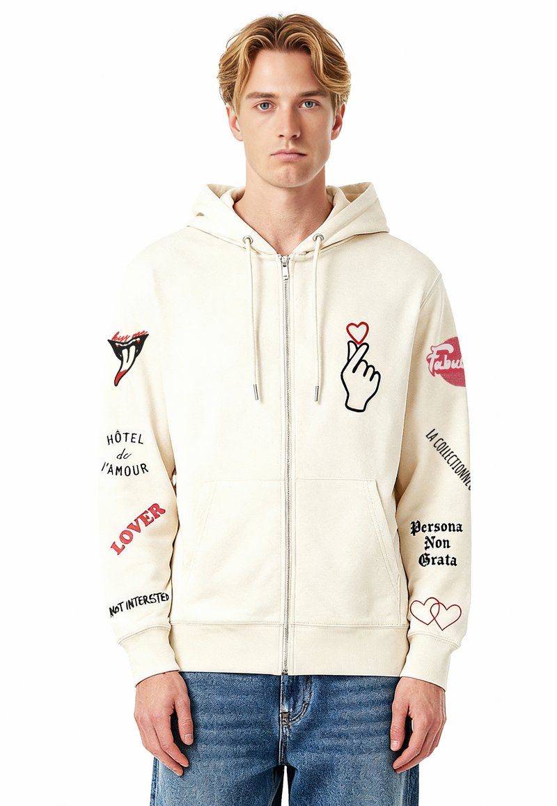 Sweat à capuche zippé en coton couleur crème avec des accents graphiques. Présente des motifs de main, de cœur et de texte en rouge et noir sur les manches et à l'avant. Poches avant incluses.