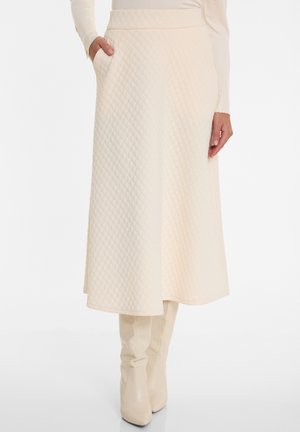 Femme portant une jupe midi matelassée crème avec poches latérales et des bottes assorties montant jusqu'aux genoux, debout devant un fond blanc.