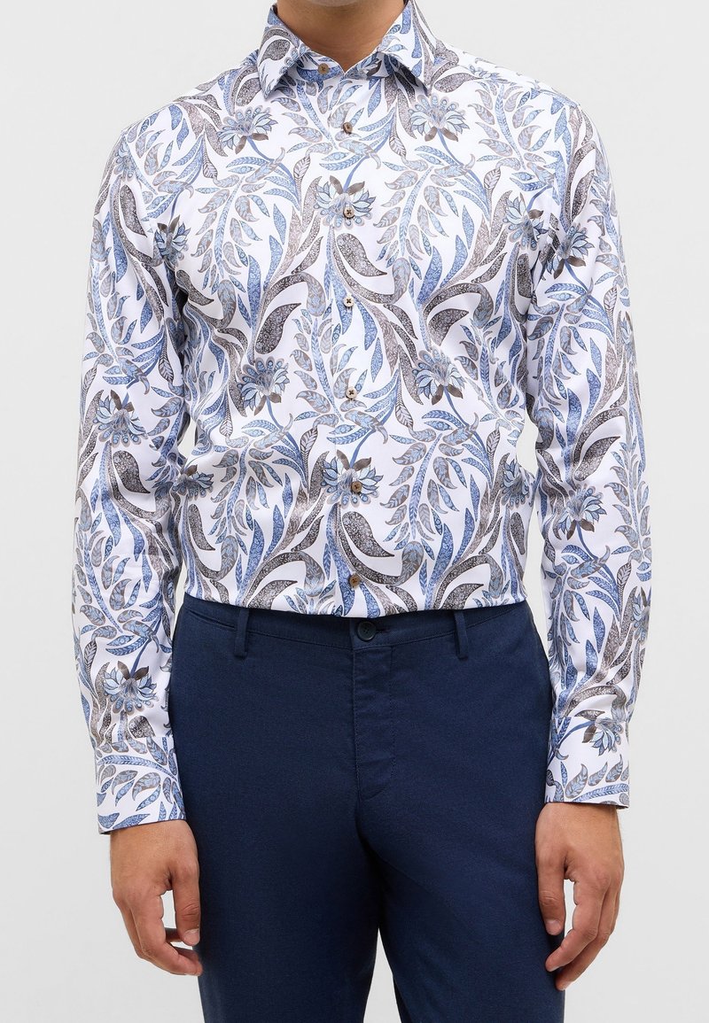 Homme portant une chemise blanche à manches longues avec des motifs floraux bleus et gris et un pantalon bleu foncé, debout, les bras détendus.