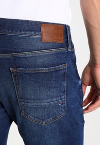 Jeans de mezclilla en azul oscuro con textura suave. Cuenta con un parche de cuero marrón en la cintura trasera y una pequeña etiqueta de marca en el bolsillo trasero.