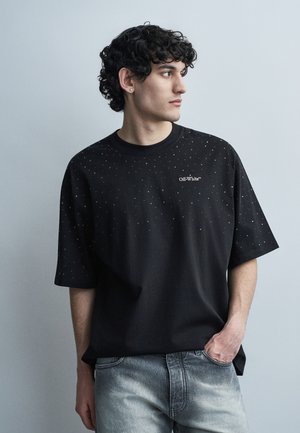 Νεαρός με σγουρά μαλλιά, που φοράει μαύρο πουκάμισο Off-White με λευκά στίγματα και ανοιχτό γκρι τζιν, κοιτάζει προς τα πλάγια με το ένα χέρι στην τσέπη.