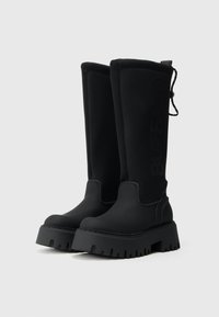 Juodi iki kelių batai, pagaminti iš lygaus medžiagos, su storu, briaunotu padu ir įspaustu logotipu šone, bei su užtrauktuku gale.