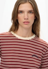 Jonge vrouw met lang bruin haar, gekleed in een rood-witte gestreepte shirt met het "Superdry"-logo op de borst, kijkt recht vooruit.