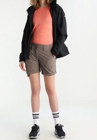 Giacca nera leggera, t-shirt arancione, pantaloni corti khaki, calze bianche con righe nere e scarpe da ginnastica nere. Stile sportivo da esterno.