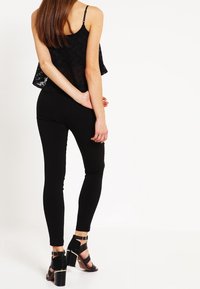 Sort blondelace crop top parret med sorte skinny bukser. Modellen har sorte hæle sandaler med ankelremme på, hvilket viser en slank silhuet.