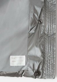 Tela de chaqueta aislante plateada con textura suave. Cuenta con una etiqueta tejida para el nombre y el teléfono, y un botón de presión para el cierre.