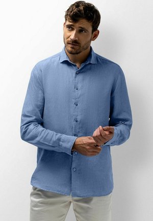 Mann mit kurzen braunen Haaren, der ein hellblaues Langarmhemd und helle Hose trägt und seine linke Manschette justiert.