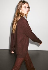 Burgundy oversized tröja med långa ärmar, mjuk textur och ribbad nederkant, kombinerad med bruna läderbyxor, stående mot en ljus bakgrund.