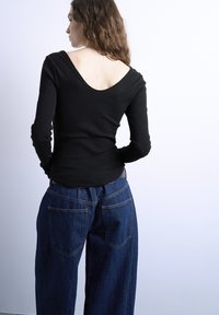 Γυναίκα με μακριά κυματιστά μαλλιά, φορώντας μαύρο μπλουζάκι με μακριά μανίκια και ευρύ ντεκολτέ στην πλάτη, καθώς και άνετο σκούρο μπλε τζιν, κοιτάζοντας προς το πλάι.