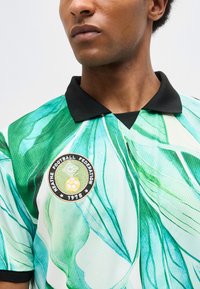 Άνδρας που φοράει ποδοσφαιρική φανέλα με πράσινο και λευκό φυλλώδες σχέδιο με μαύρο γιακά και το έμβλημα «Weathe Football Federation 1978».