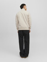 Beige zip-up sweatshirt med høj krave, afslappet pasform og ribbede manchetter, parret med sorte løse jeans og brune sko.