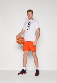 Baltas NBA logotipas marškinėliai, oranžinės sportinės šortai, laiko krepšinį, vilkintis juodus ir raudonus sportinius batelius, stovintis ant vientisos fono.