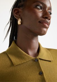 Olivengrøn strikket polo-shirt med krave og tre mørke knapper. Modellen bærer en gulddråbeørering, og hendes hår er stylet i fletninger.