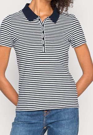 Frau trägt ein marineblau-weiß gestreiftes Poloshirt mit kurzen Ärmeln und dunklem Kragen, dazu blaue Jeans, steht mit den Händen hinter dem Rücken.