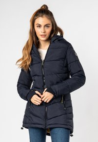 Jonge vrouw met lang haar die een gedessineerde, donkergroene gewatteerde jas met capuchon en blauwe spijkerbroek draagt, tegen een effen achtergrond.