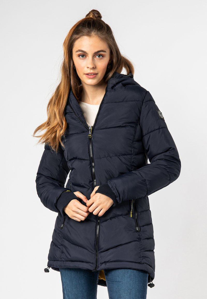 Jonge vrouw met lang haar die een gedessineerde, donkergroene gewatteerde jas met capuchon en blauwe spijkerbroek draagt, tegen een effen achtergrond.