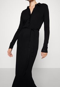 Robe noire longue et ajustée avec un col, une patte de boutonnage, des manches longues et une taille nouée, confectionnée dans un tissu doux et lisse.