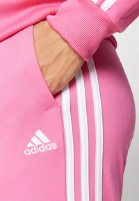 Rozā Adidas sporta bikses ar ribotu jostu, kurām ir baltas trīs svītras un izšūta logo kreisajā gurnu kabatā.
