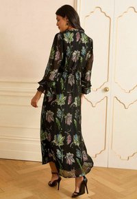 Schwarz langärmliges Kleid mit floralen Mustern in Grün, Blau und Lila. Durchsichtiges Material, geraffte Taille, Rüschensaum und fließende Silhouette.