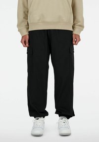 Pantalons cargo noirs avec poches latérales, poignets élastiques et coupe décontractée, associés à des baskets blanches et un sweat-shirt beige.