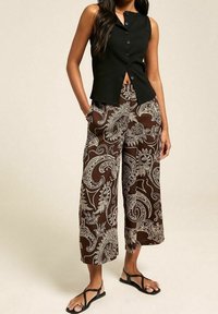 Černý bezrukávový top s předními knoflíky, v kombinaci s hnědými kalhotami s paisley vzorem a širokými nohavicemi. Modelka má na sobě černé žabky.
