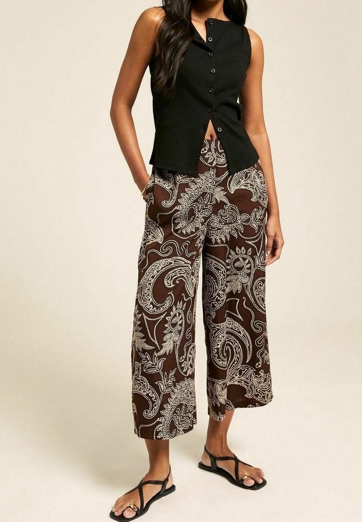 Černý bezrukávový top s předními knoflíky, v kombinaci s hnědými kalhotami s paisley vzorem a širokými nohavicemi. Modelka má na sobě černé žabky.