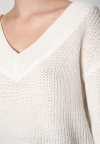 Cremfarbener Strickpullover mit weitem V-Ausschnitt, geripptem Kragen und einem strukturierten Muster mit offenen Maschen. Weiches, leichtes Material.