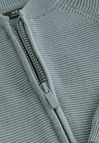 Krupni plan djelomično zatvorenog sivog rebrastog pletiva s plastičnim patentnim zatvaračem i tkanom etiketom unutar ovratnika.