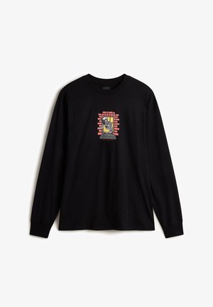 Černé tričko s dlouhým rukávem vyrobené z bavlny, které na přední straně zobrazují centrální grafiku kostry orámovanou žlutou barvou na pozadí červených cihlových zdí.