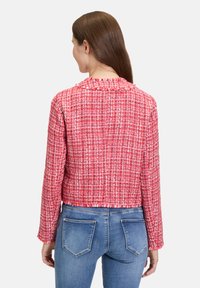 Czerwona tweedowa kurtka o teksturowanym wykończeniu i postrzępionym kołnierzyku, z długimi rękawami i skróconym fasonem. Połączona z niebieskimi dżinsami.