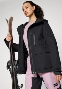 Melnā slēpošanas jaka ar vairākiem kabatām, priekšējais rāvējslēdzējs un teksturēta kapuce. Modelis tur slēpes, valkājot rozā augšu un bikses.