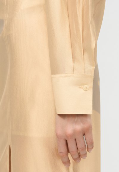 Χέρι που ακουμπά στο πλάι, φοράει μπεζ ρούχο με μακριά μανίκια, μανσέτα με κουμπί και διακριτική υφή υφάσματος.