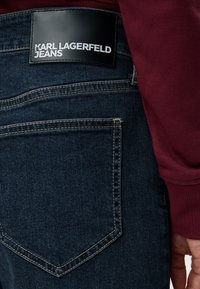 Temno modri jeans iz denima z vidno žepom. Na pasu je črno usnjeno oznako z belim napisom "KARL LAGERFELD JEANS."