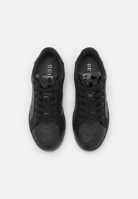 Baskets noires avec une surface texturée, présentant des logos en relief, un bout rond, et des lacets noirs. Accents brillants près de la languette.