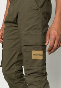 Olivegröna cargobyxor i bomull, med flera fickor, en texturerad yta och ett märke som säger "DENIM PROJECT."